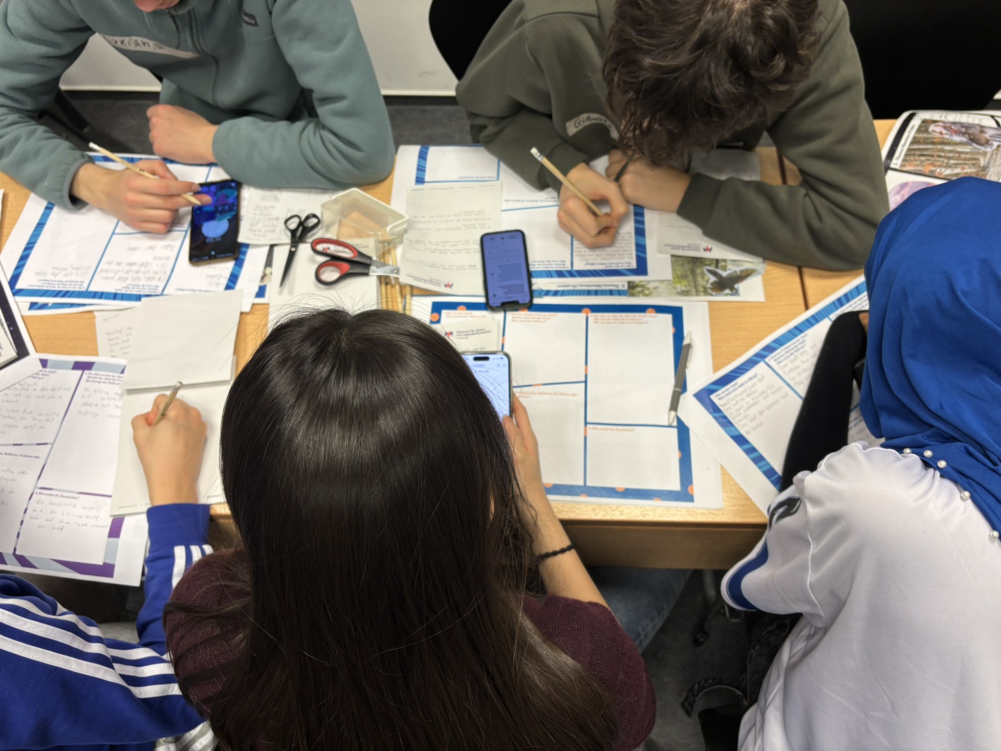 Mehrere Jugendliche sitzen gemeinsam an einem Tisch und schreiben auf Arbeitsblätter. Smartphones, Stifte und Unterrichtsmaterialien liegen auf dem Tisch. Die Szene zeigt konzentrierte Gruppenarbeit im Unterricht.