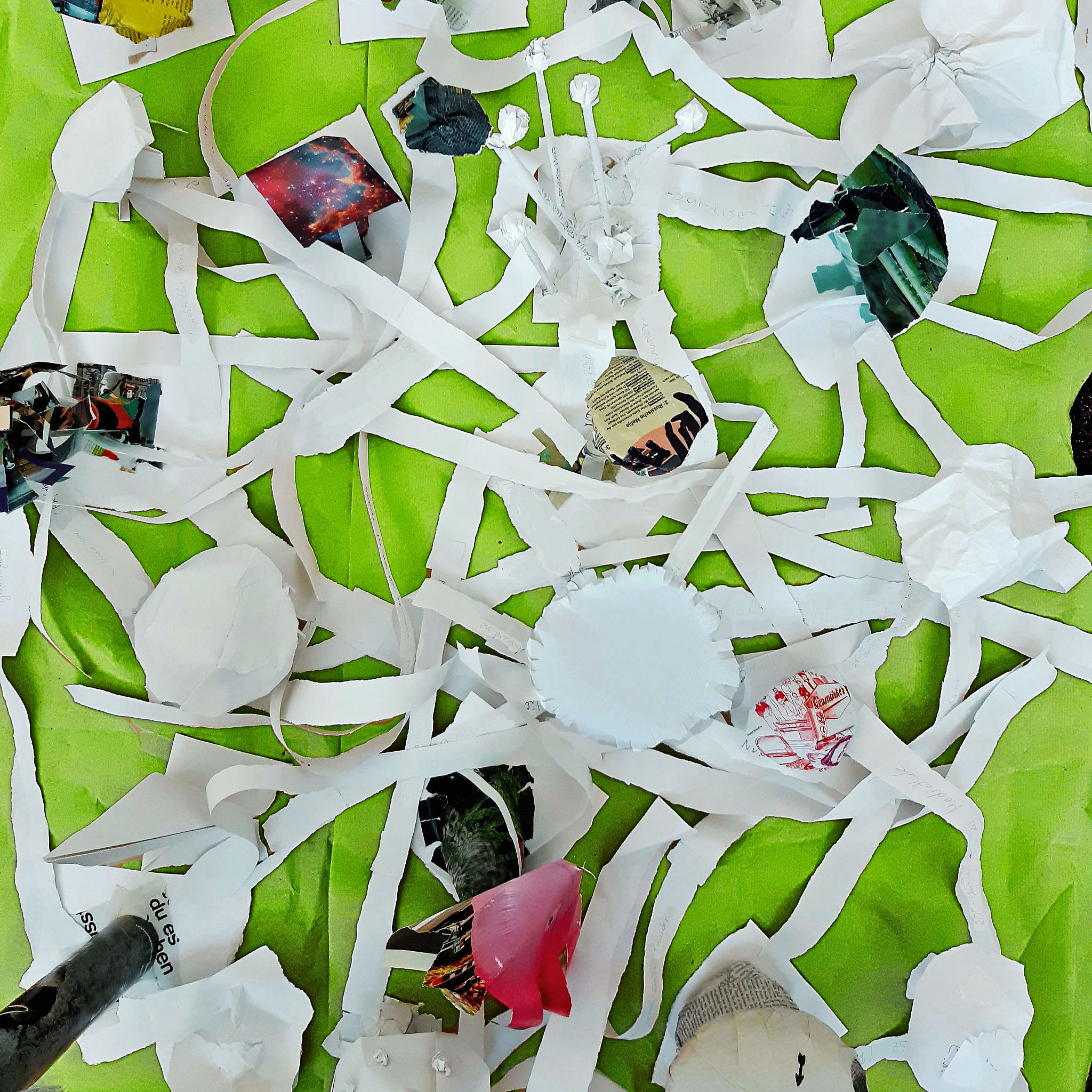 Künstlerische Papierinstallation auf leuchtend grünem Untergrund. Zahlreiche lange, weiße Papierstreifen verbinden runde, gefaltete und zerknüllte Papierformen zu einem dichten, netzartigen Geflecht. Einige Elemente bestehen aus Zeitungsausschnitten und farbigen Bildern. Die Arbeit erinnert in ihrer verzweigten Struktur an ein Pilzmyzel oder ein organisches Netzwerk.